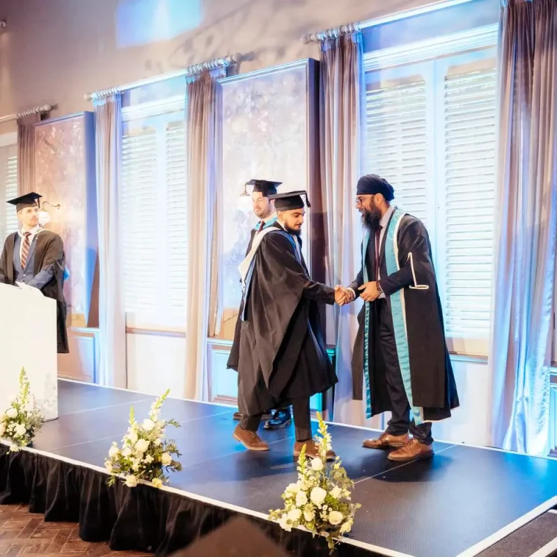 Graduates in caps and gowns shaking hands on a stage with a podium and floral arrangements, under soft lighting.