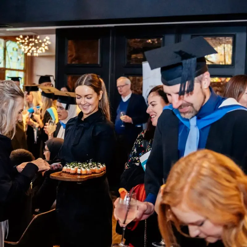 Graduates in academic gowns and caps celebrating with canapés and drinks in a warmly lit room.