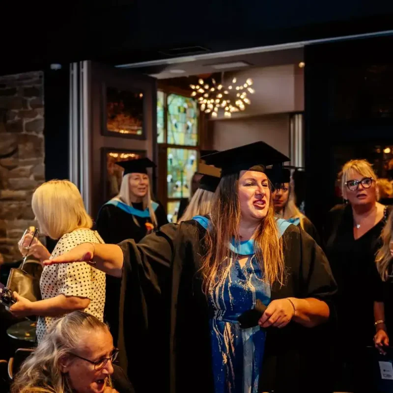Graduates in black gowns and caps celebrate indoors, holding certificates and glasses, with a stone wall and chandelier in the background.