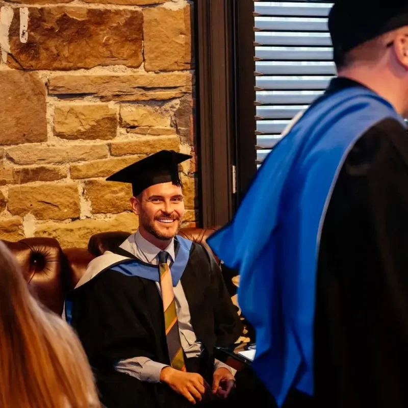 A man in graduation gown and cap smiling, seated in front of a stone wall, during a graduation ceremony.