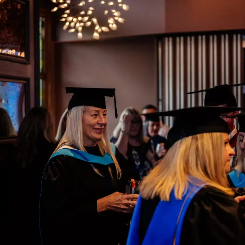Graduates in academic gowns and caps celebrating in a warmly lit room with framed artwork and a chandelier.