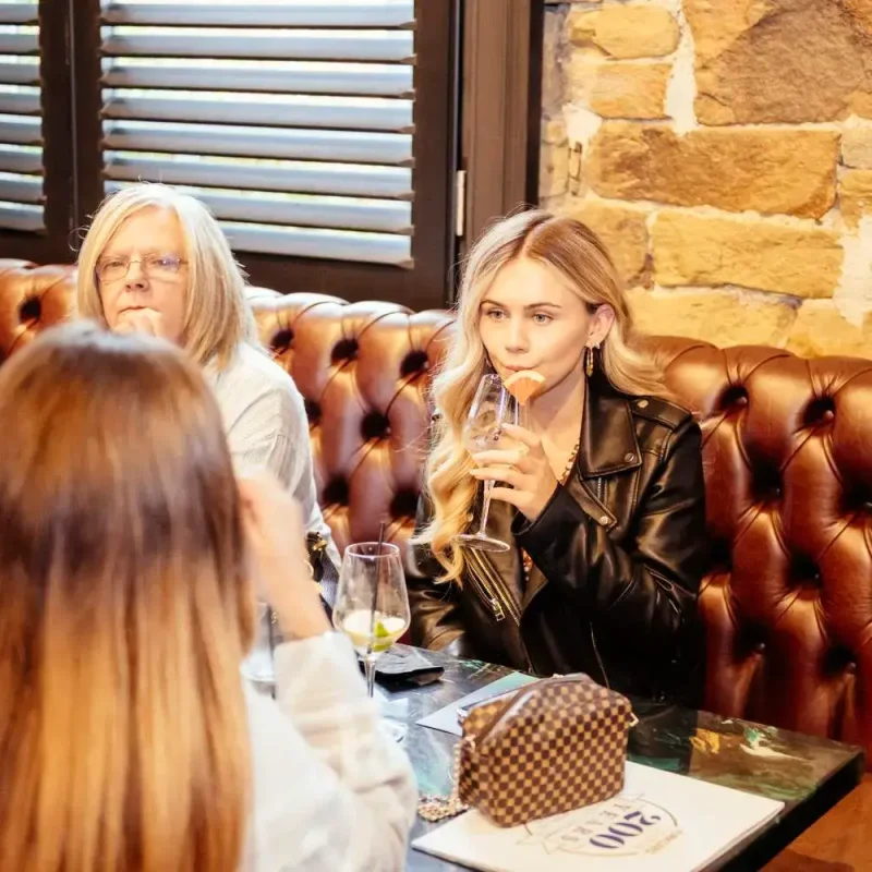 Three women in a pub booth, one sipping wine, another on her phone, and a third listening. Stone wall and wooden shutters in the background.