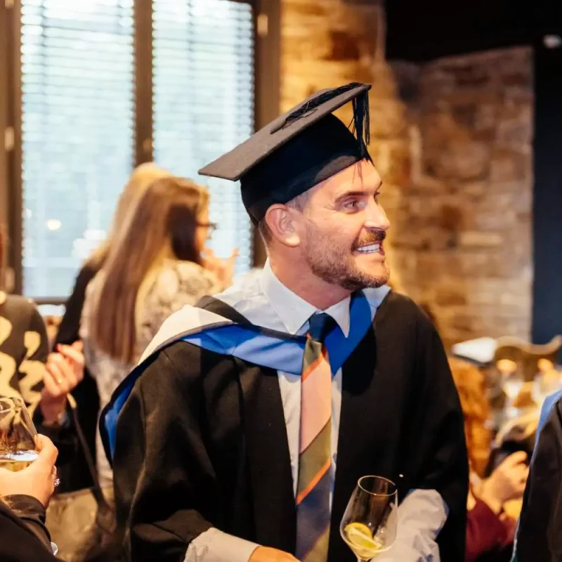 Graduates in academic gowns and caps celebrating with drinks in a warmly lit room, marking their academic achievements.