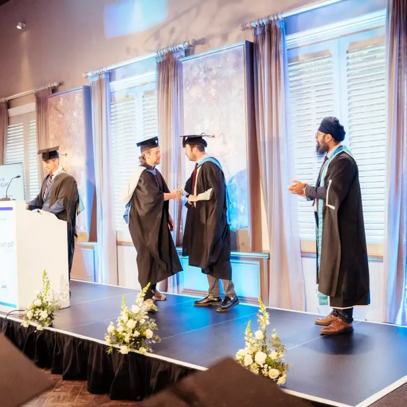 Graduates in caps and gowns receive certificates on stage with a podium and floral arrangements, under soft lighting and elegant decor.