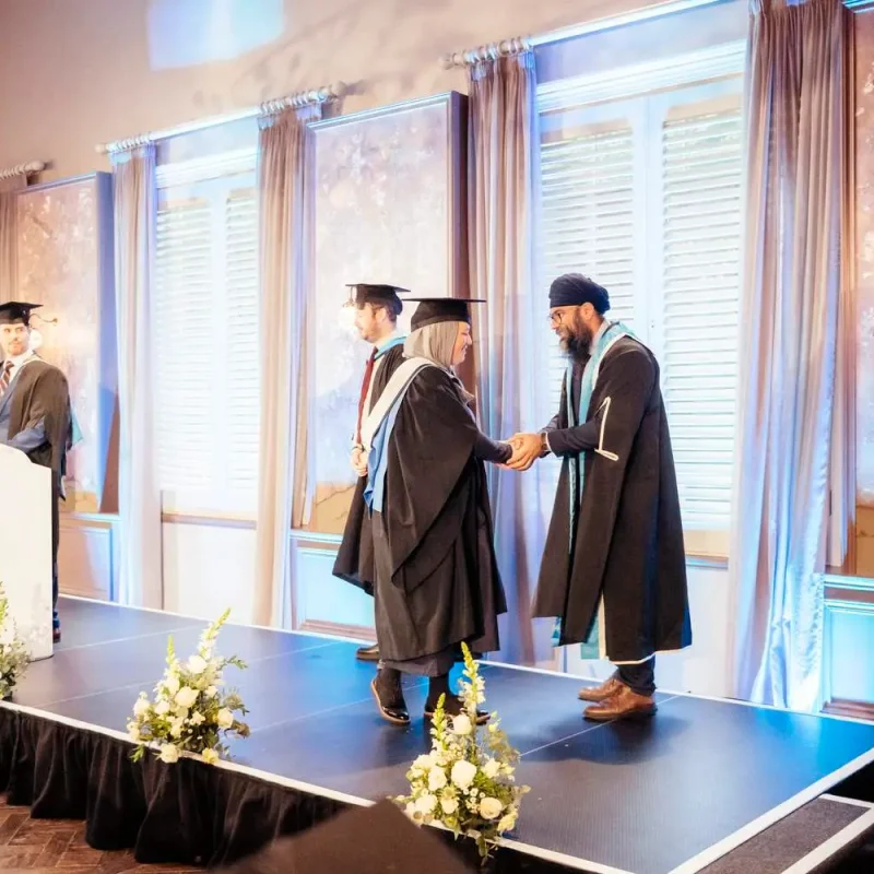 Graduates in academic gowns receiving certificates on a stage with floral arrangements and a podium.
