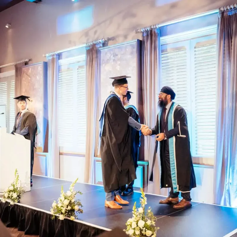 Graduation ceremony with two individuals shaking hands on stage, surrounded by floral arrangements and a podium with a university logo.