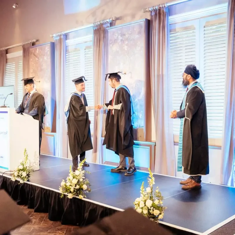 Graduation ceremony with graduates in gowns receiving certificates on stage, flanked by academic staff and floral decorations.