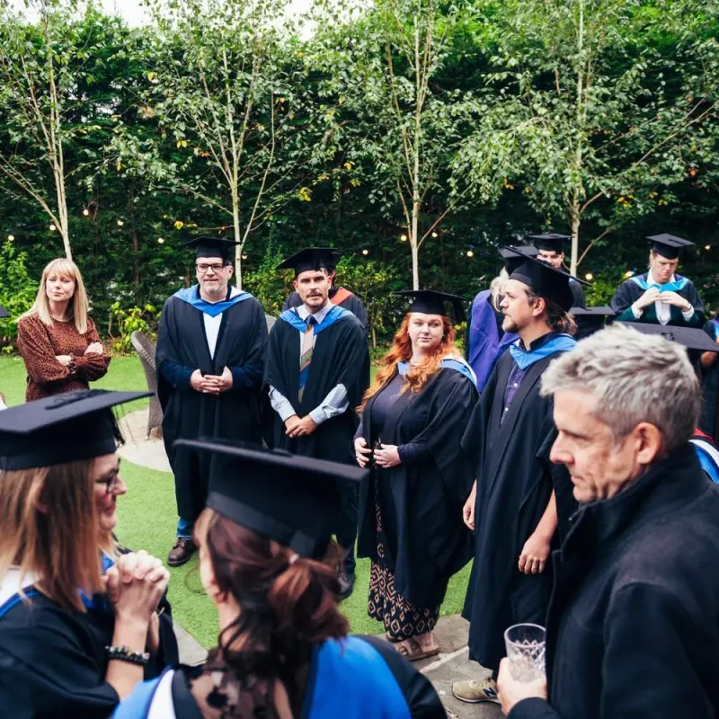 Graduates in black gowns and caps gather outdoors with faculty, celebrating academic achievements amidst lush greenery and string lights.