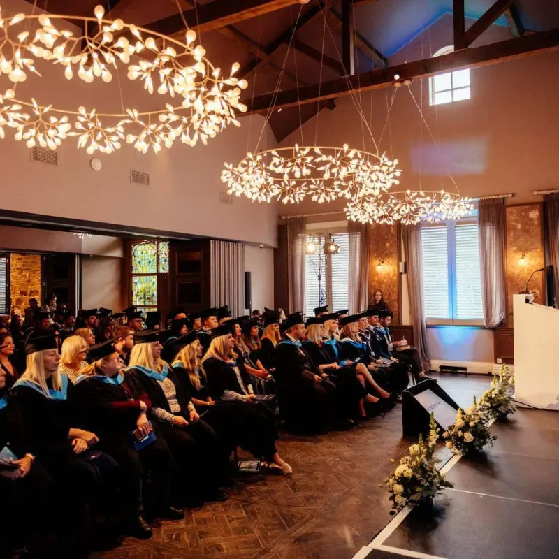 Graduates in caps and gowns seated in rows, facing a speaker at a podium in a warmly lit hall with ornate chandeliers.