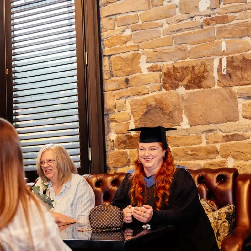 A woman in graduation gown and cap smiling with two women at a table in a stone-walled room.