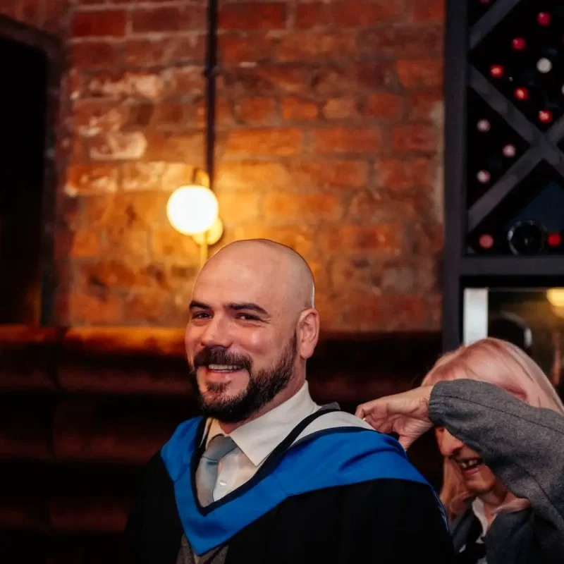 A man in academic regalia smiling, with a woman adjusting his hood in a warmly lit room with brick walls and wine bottles.