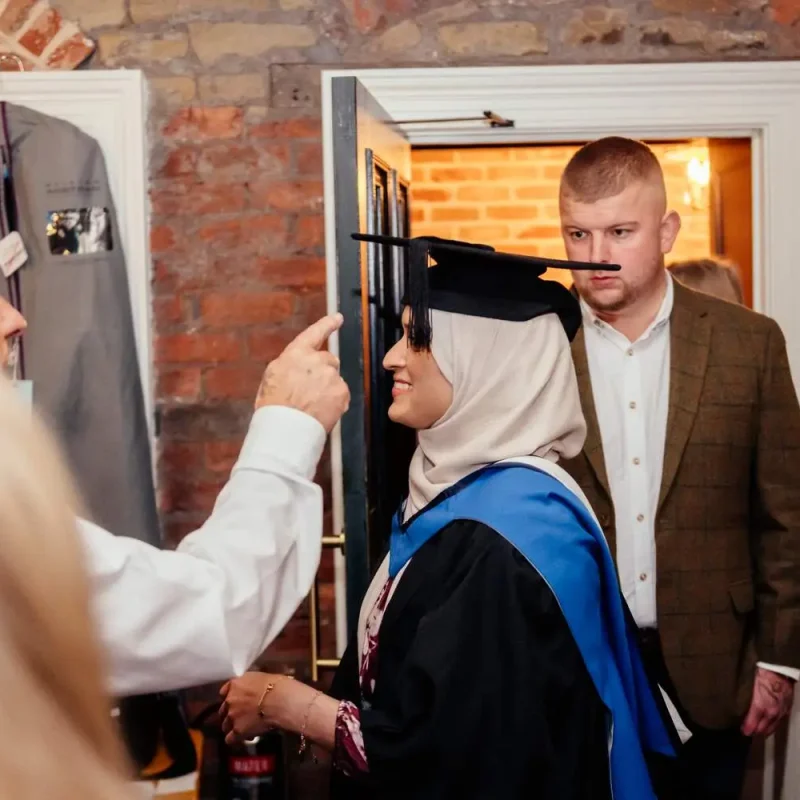 A graduate in academic regalia stands in a doorway, smiling as an older man playfully points at a moustache drawn on their face.