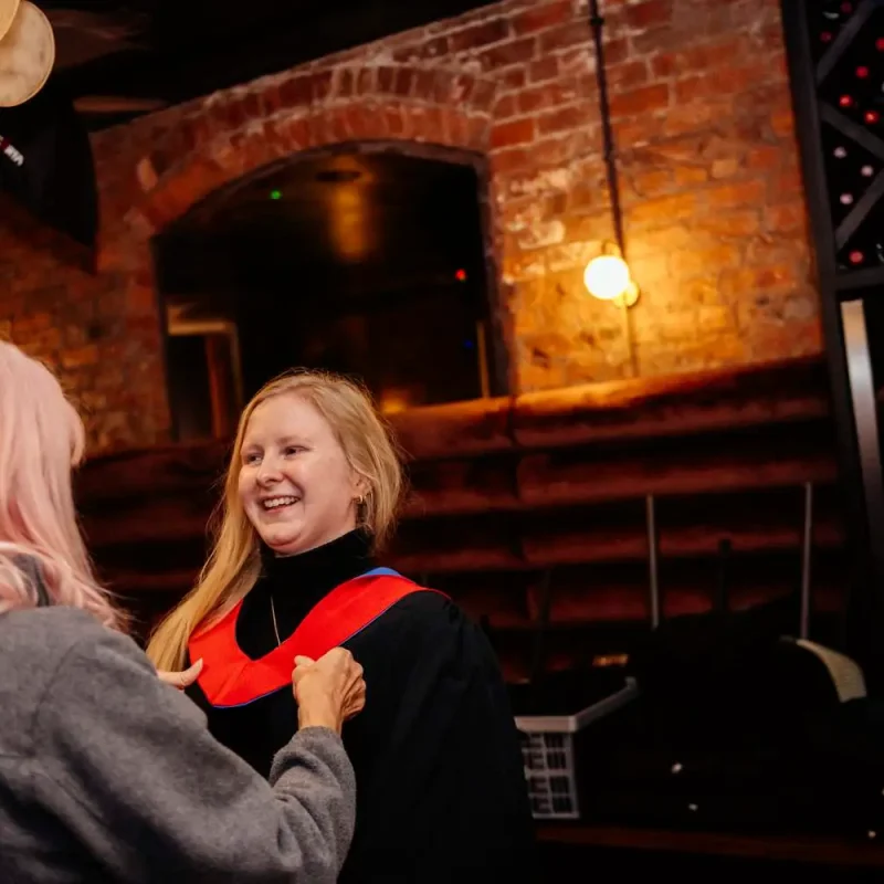 Two women in academic gowns, one smiling, in a brick-walled room with hanging lights and a wine rack, celebrating a graduation achievement.