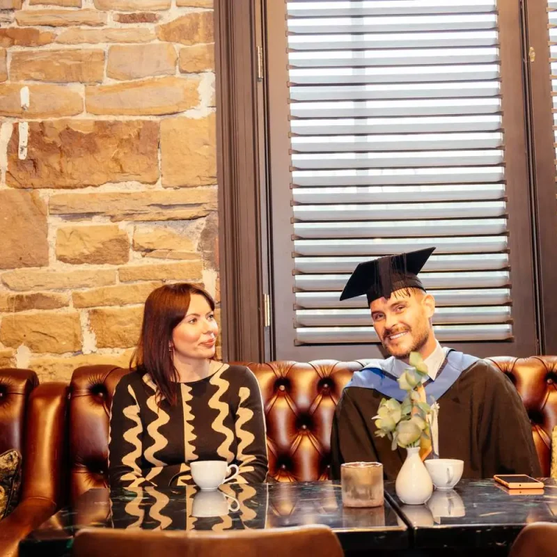 Two graduates in academic regalia sit on a brown leather sofa, smiling and chatting over coffee in a stone-walled room.