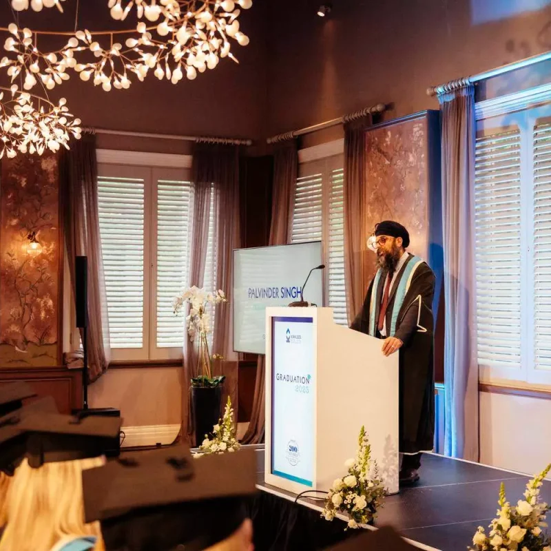 A person in academic regalia stands at a podium in a room with ornate decor, addressing graduates seated at tables.