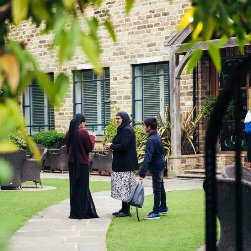 Students in academic gowns conversing outside a historic stone building with lush greenery and outdoor seating.