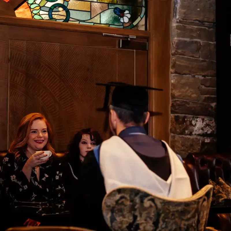 A graduate in academic regalia converses with two women in a cosy, wood-panelled room with stained glass windows, suggesting a celebratory academic event.