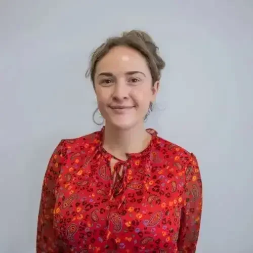 A woman in a red paisley blouse smiles against a plain background, exuding a welcoming and approachable demeanour, ideal for an educational setting.
