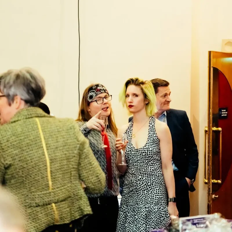 A group of people in formal attire, including a woman in a polka dot dress, engaged in conversation at a college event.