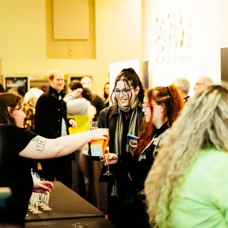 A group of people in a gallery, some holding drinks, engaged in conversation near a table with champagne flutes.