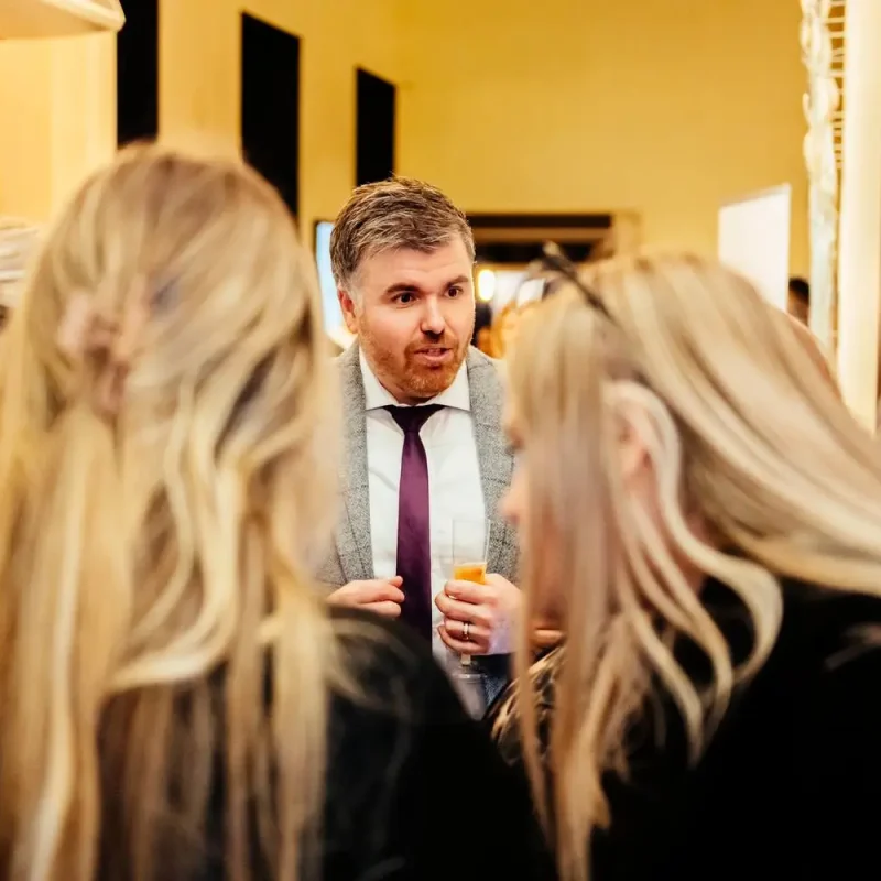 A man in a suit talking to three women in a gallery with framed portraits.