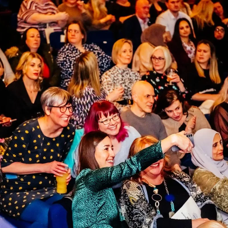 A diverse group of people seated in an auditorium, smiling and engaged, possibly attending a lecture or seminar.