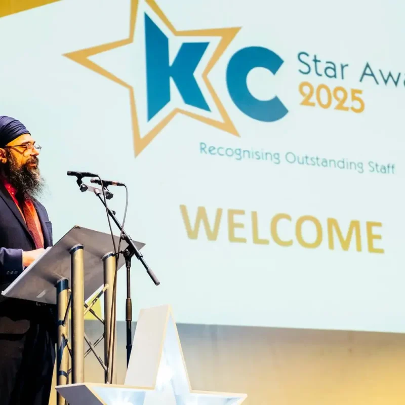 A man in a suit stands at a podium in front of a projection screen displaying 'KC Star Awards 2025' and 'Recognising Outstanding Staff'.