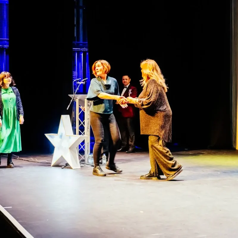 Two women shaking hands on a stage with a star prop, watched by others in formal attire. College event setting.