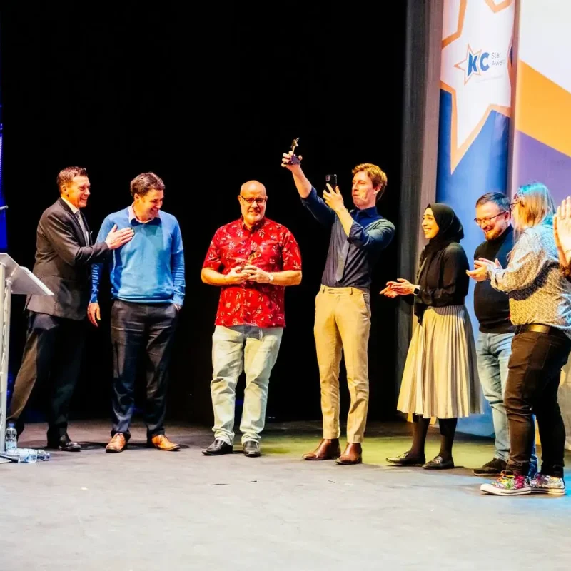 A group of people on a stage, some clapping and one taking a photo, during an award ceremony.