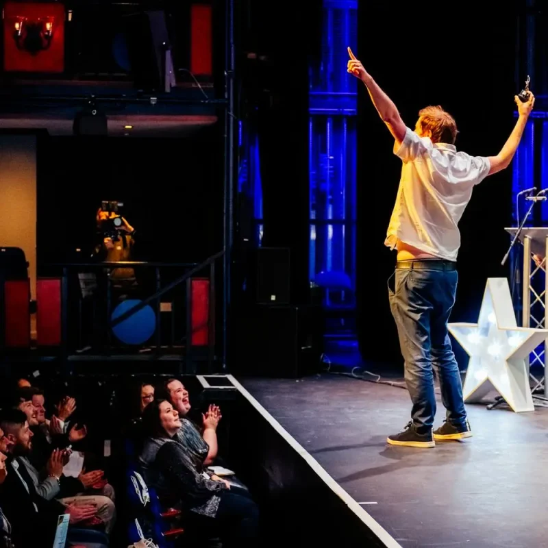 A man on stage raises his arms in celebration, surrounded by applauding audience members and a large illuminated star.