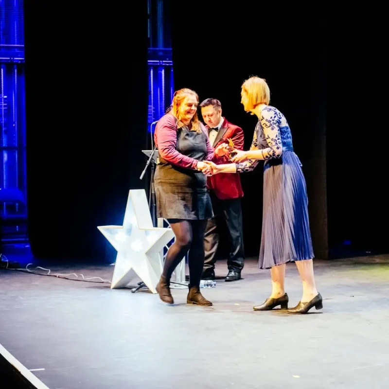 Three people on a stage, two women and a man in a suit, holding hands and smiling, with a large white star in the background.