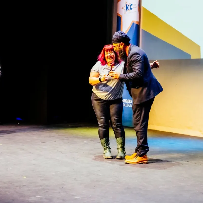 A woman in a grey top and green boots receives an award on stage from a man in a suit, both smiling. A man in a suit stands at a podium in the background.