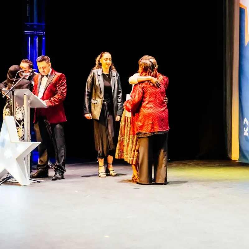 Four people on stage at a college awards ceremony. Two are hugging, one in a red jacket, and two others stand near a podium with a star design.