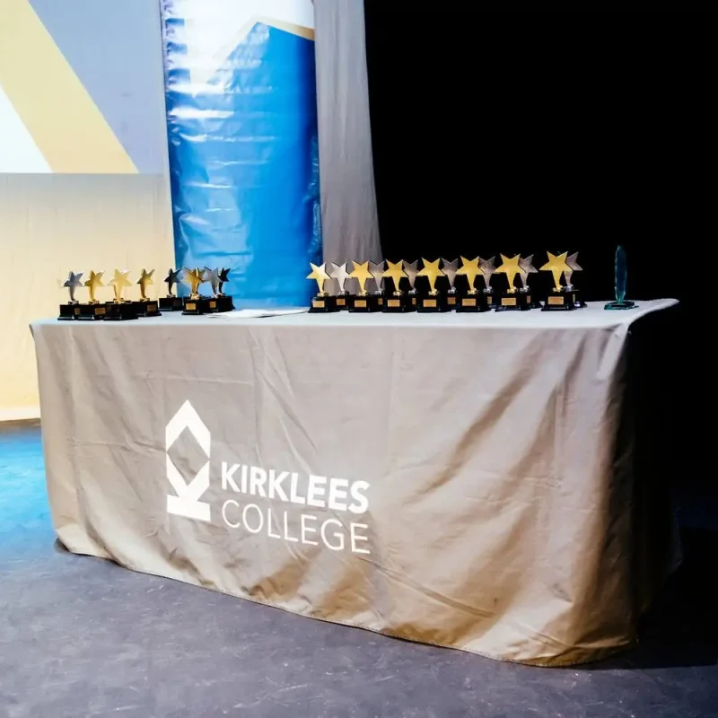 Kirklees College awards ceremony table with gold star trophies and a blue banner backdrop.