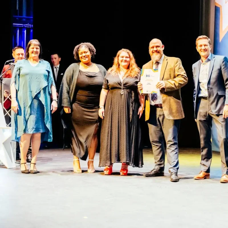 Five people on a stage, one holding a certificate, smiling and dressed in formal attire. A large illuminated star is visible to the left.