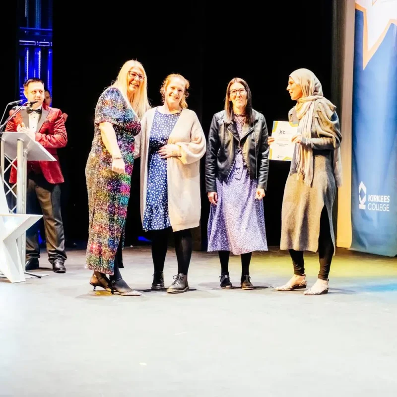 A group of four women and a man in formal attire on a stage, with one woman holding a certificate, at a college event.