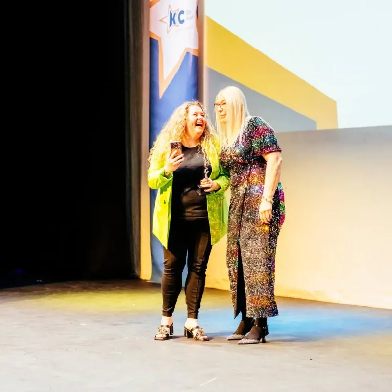 Two women on stage, one holding a phone, both smiling and engaged in conversation, likely at a college event.