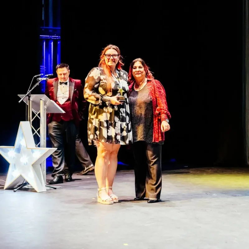 Two women holding awards on a stage with a star prop and a man in a red jacket behind a podium.