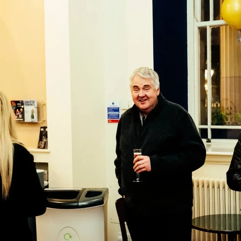 Three people in a room with balloons, one holding a drink, suggesting a social or educational event.