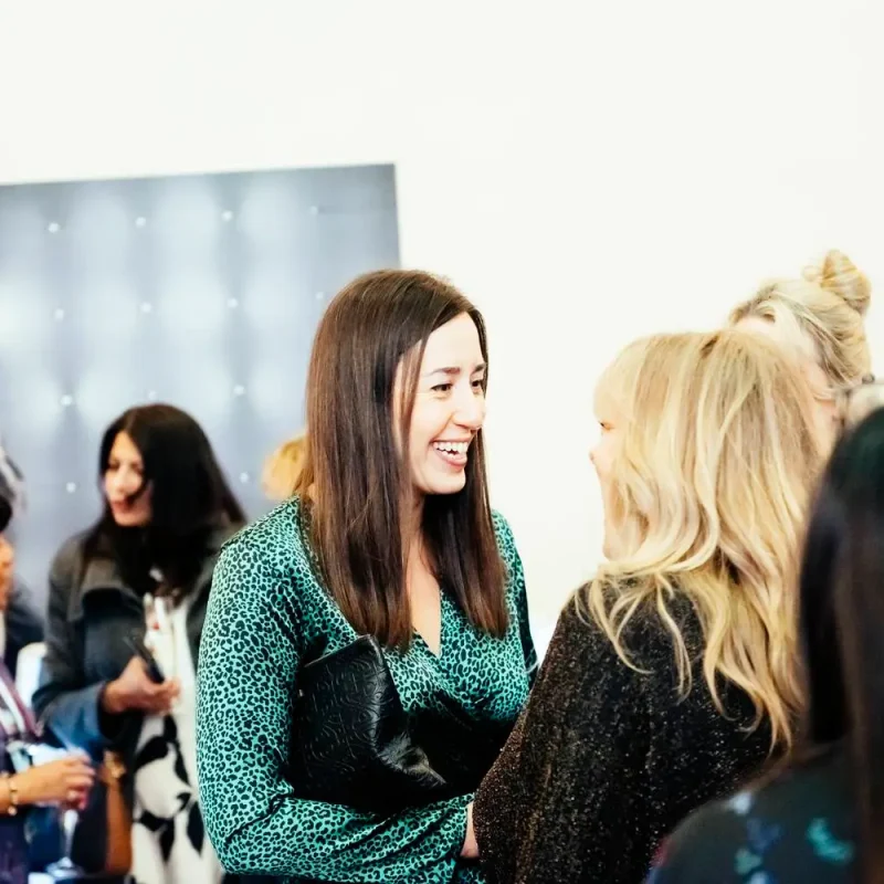 A group of women in a brightly lit room, engaged in conversation. One woman is smiling broadly, holding a black clutch. A poster of a woman in a white dress is in the background.