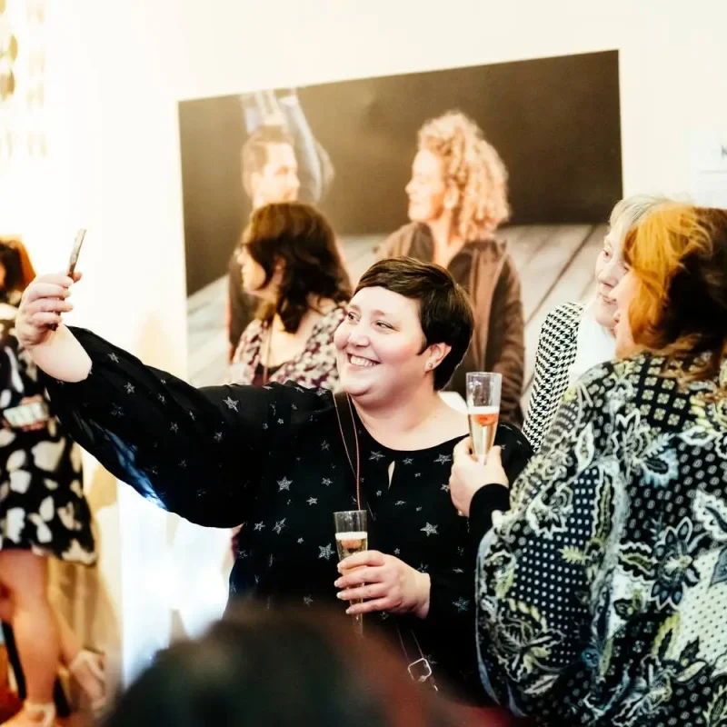 A woman in a black star-patterned top takes a selfie with two women holding drinks at a college art exhibition.