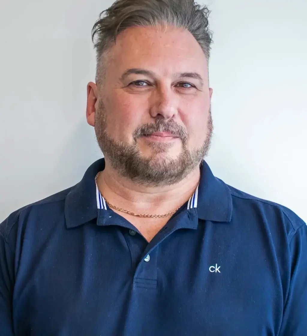 A man with a beard wearing a navy polo shirt with a white logo and a gold chain necklace, standing against a white background.