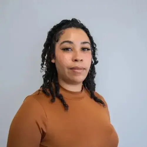 A person with curly hair wearing a brown top, standing against a grey background, smiling slightly.