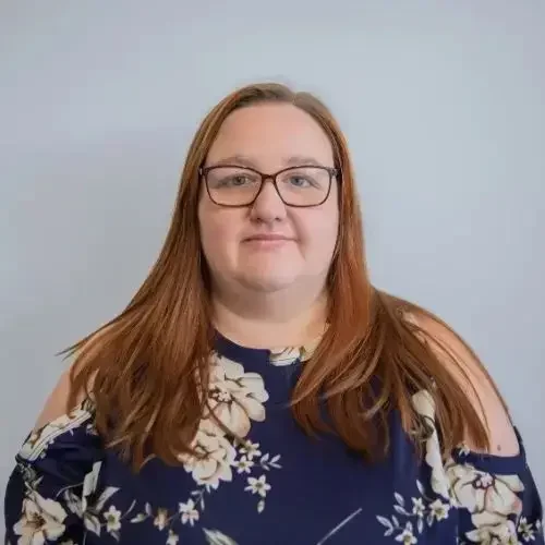 A woman with long red hair and glasses wearing a blue floral top poses for a portrait against a grey background.